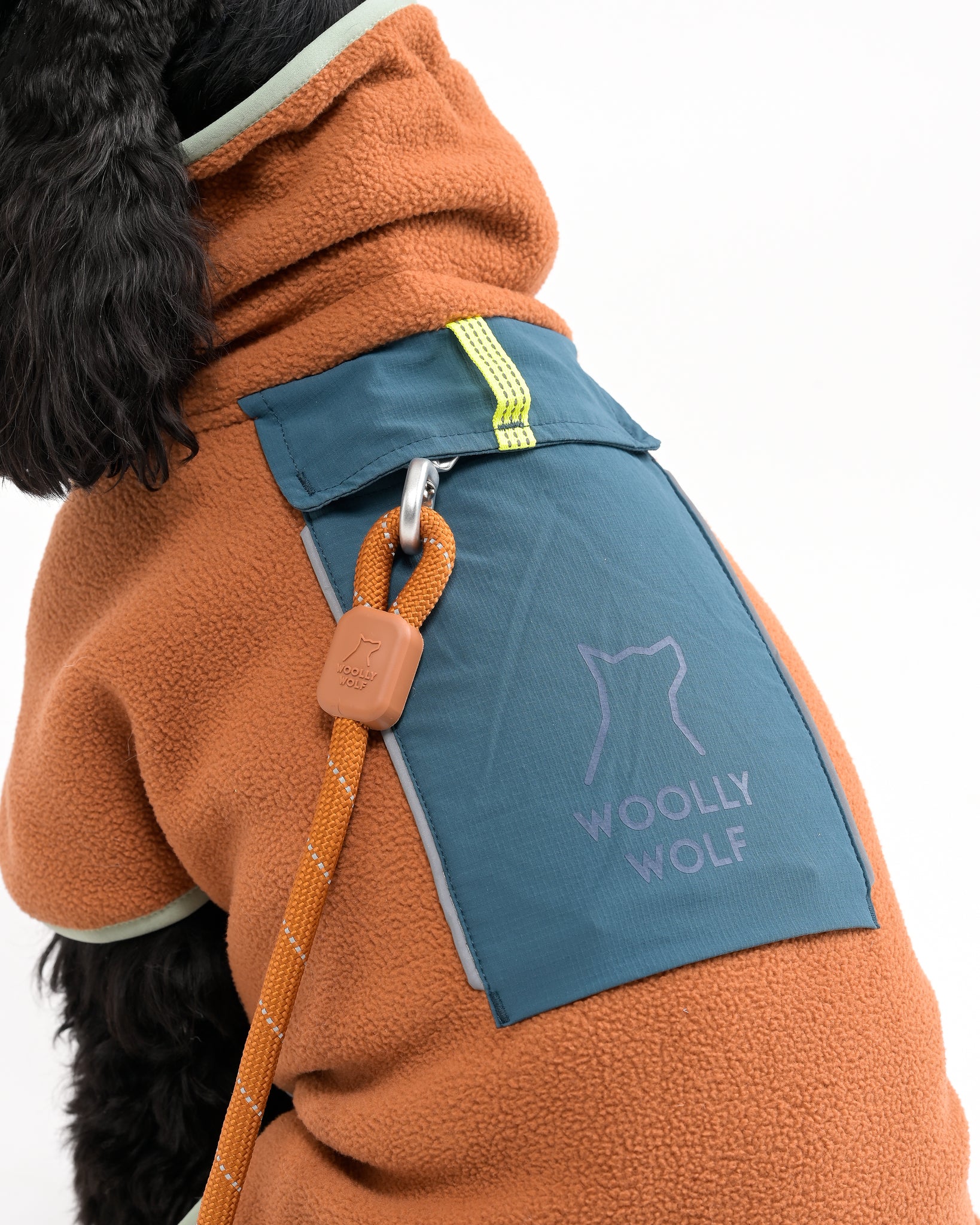 A dog wearing a brown fleece jacket with a blue pocket labeled WOOLLY WOLF and a leash attached to a metal ring on the jacket. The image is cropped to show only part of the dogs body.