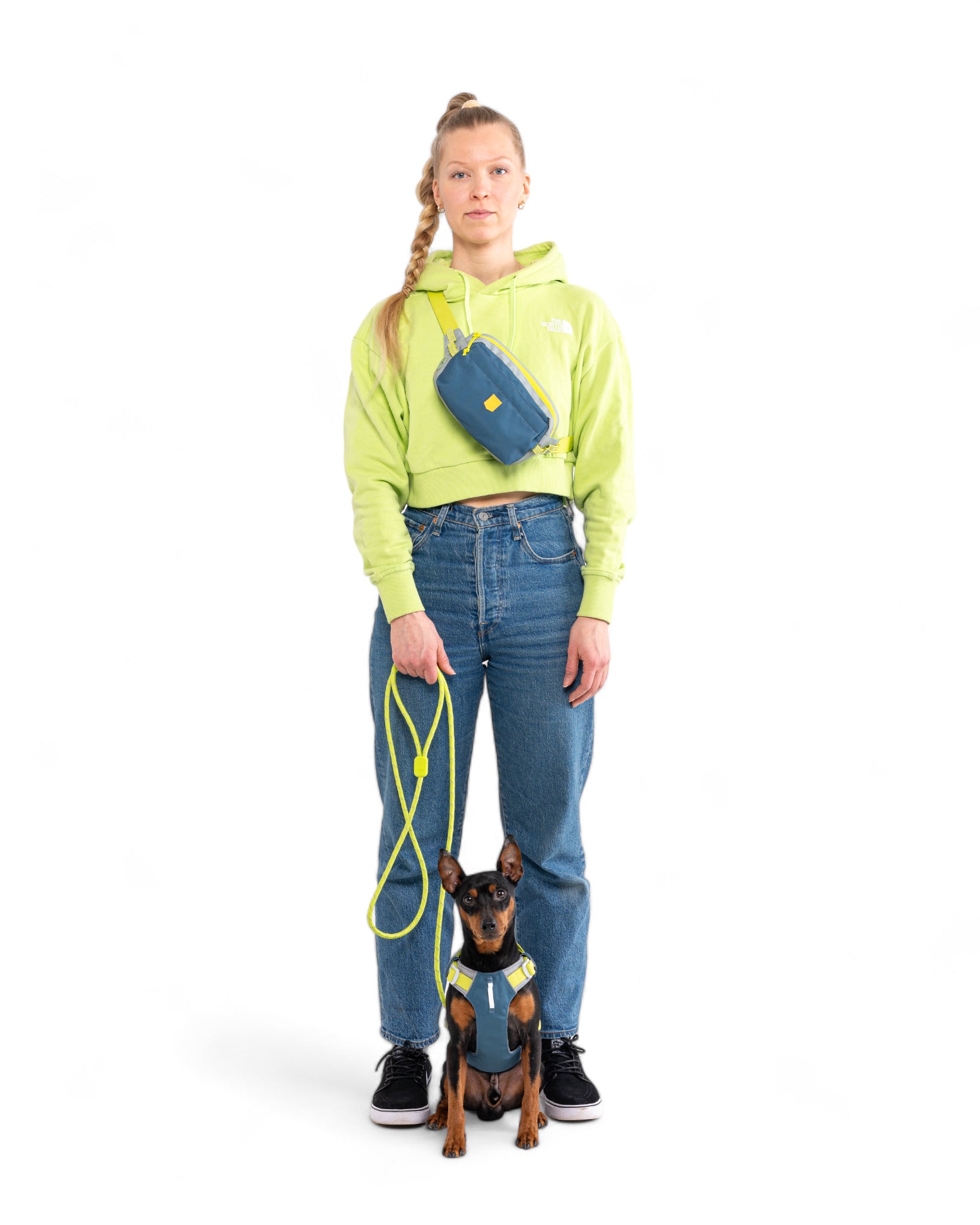 A woman in a neon green hoodie and blue jeans holds the leash of a small black and brown dog wearing the Alpha 360 Dog Harness in Deep Teal. She has a blue crossbody bag, and both face the camera against a white background.