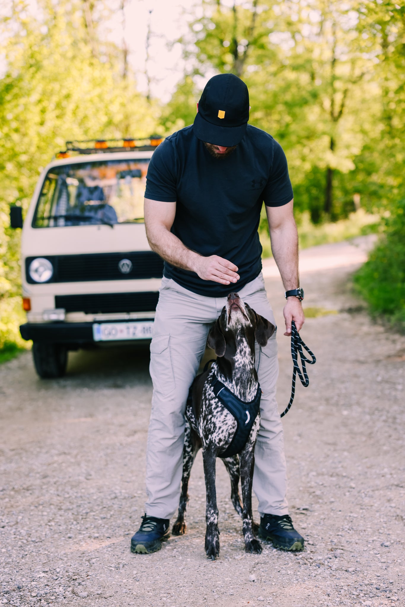 A man wearing an Alpha 360 Tech Cap Black and a t-shirt stands on a dirt path, giving a treat to a black and white harnessed dog. A white van is parked behind them among green trees.