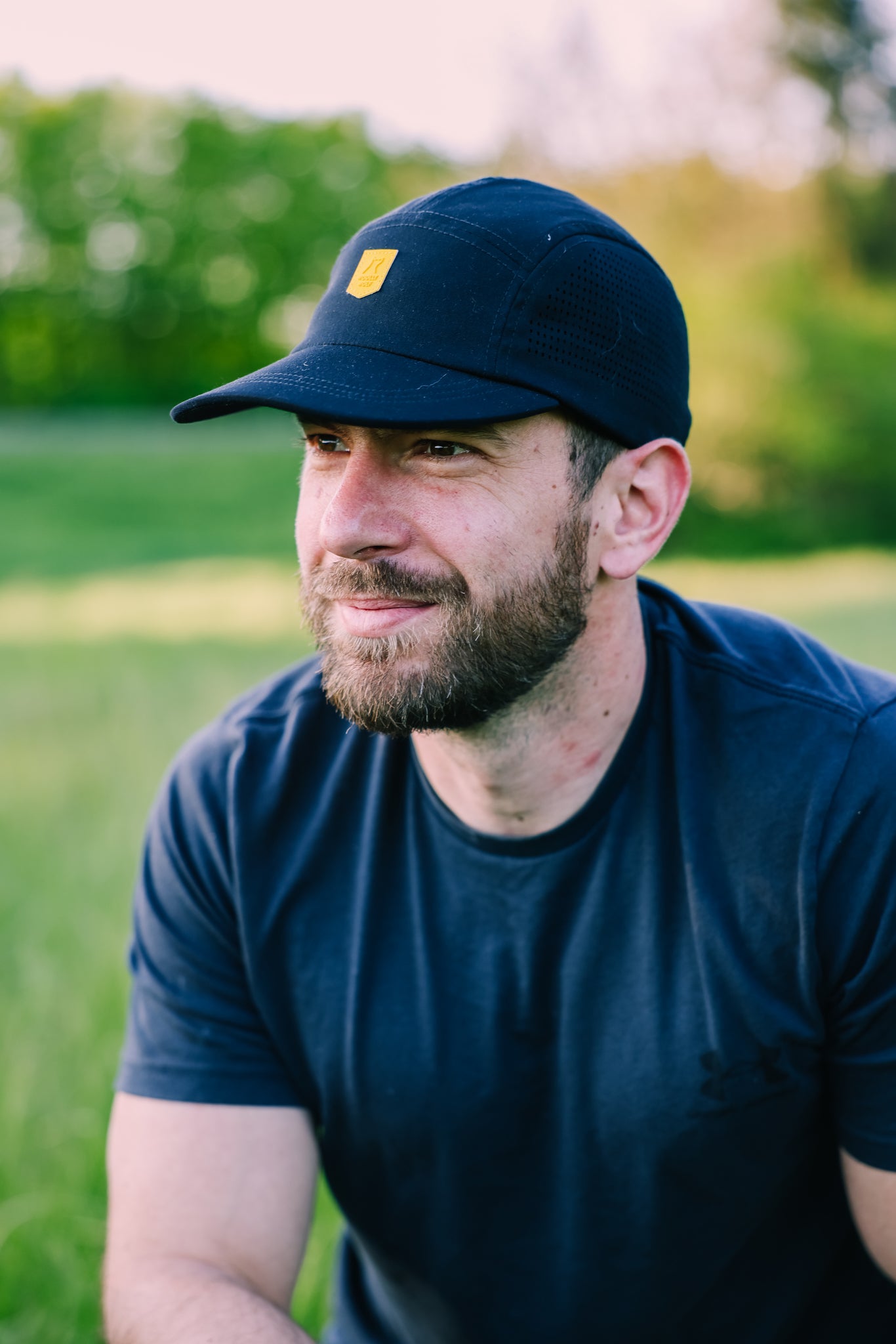 A bearded man wearing the Alpha 360 Tech Cap Black and a navy t-shirt sits on a grassy field outdoors, with trees and sunlight in the blurred background.