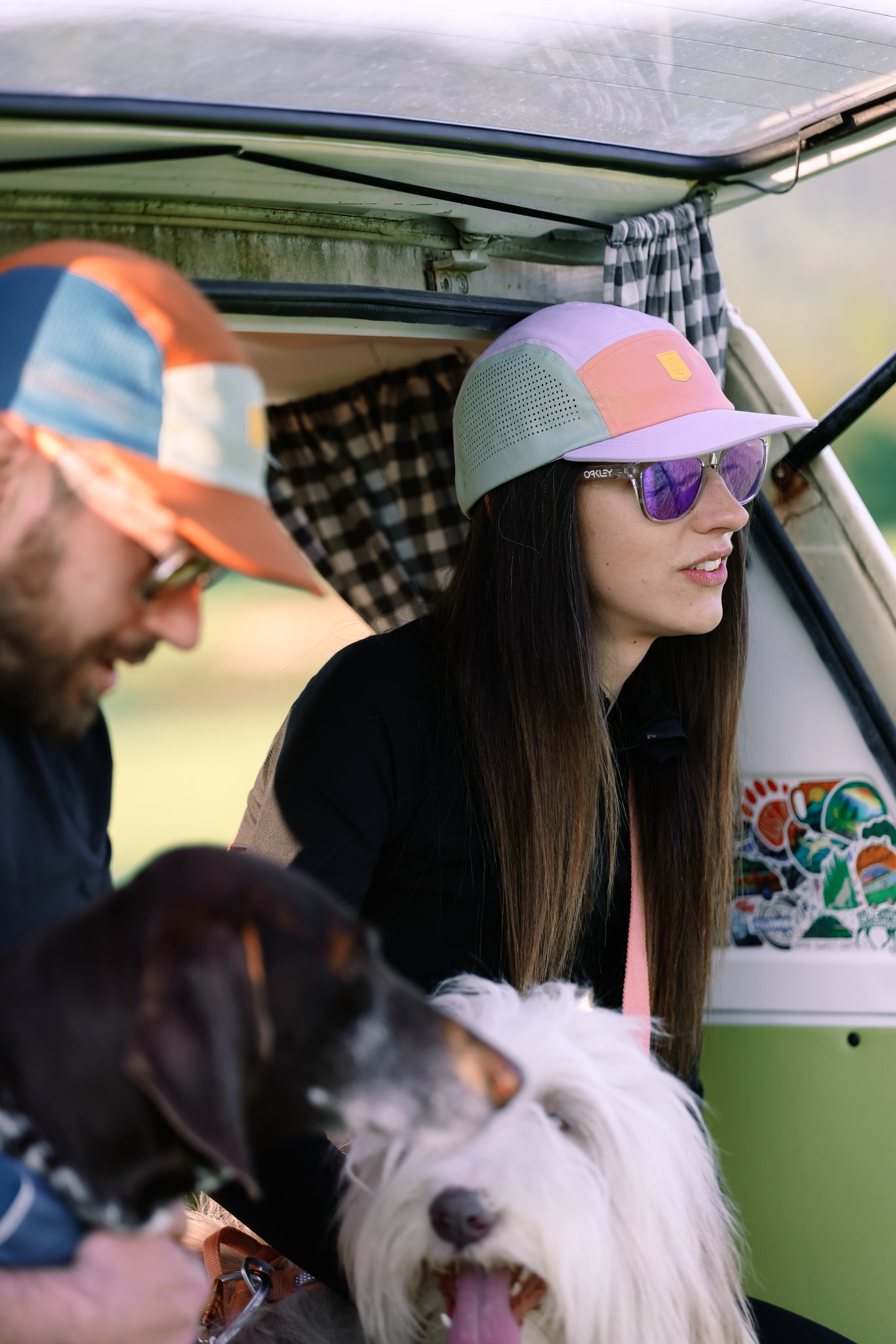 Two people relax in an open van with two dogs. The woman, in reflective sunglasses and an Alpha 360 Tech Cap Lavender, looks ahead as the man holds a dog. Their colorful hats and setting suggest a laid-back outdoor adventure.
