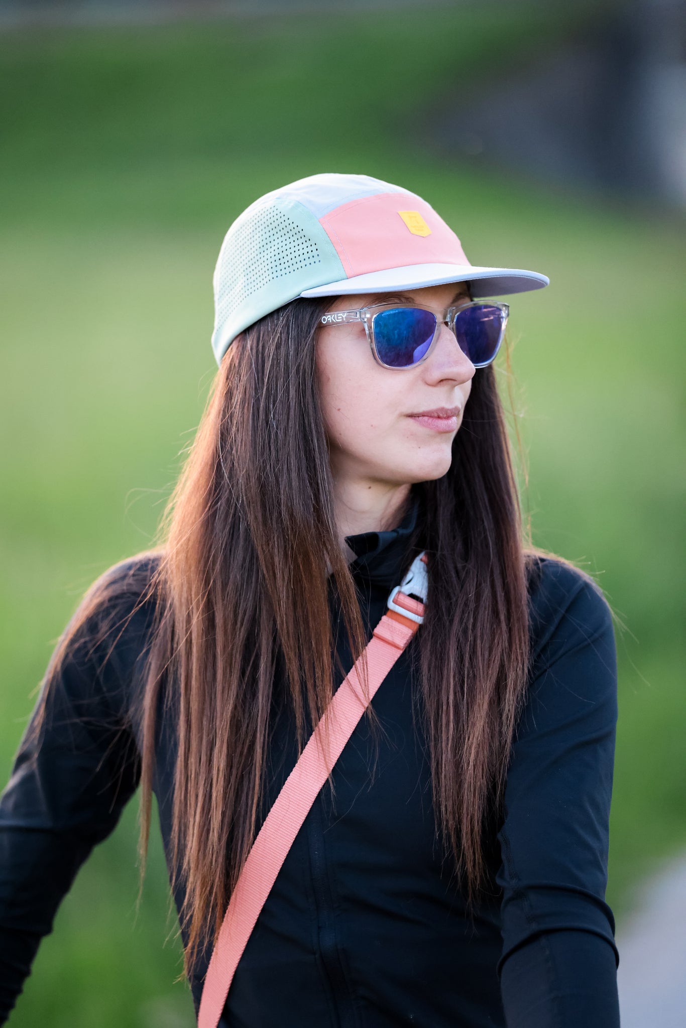 A woman with long brown hair wears the Alpha 360 Tech Cap in lavender, blue sunglasses, a black jacket, and a pink shoulder strap while standing outdoors against a blurred green grass background.
