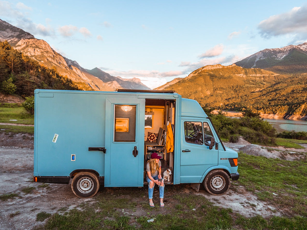 A blue van parked by mountains and a lake at sunset; a person sits on the doorstep with a dog, surrounded by peaceful, scenic nature.