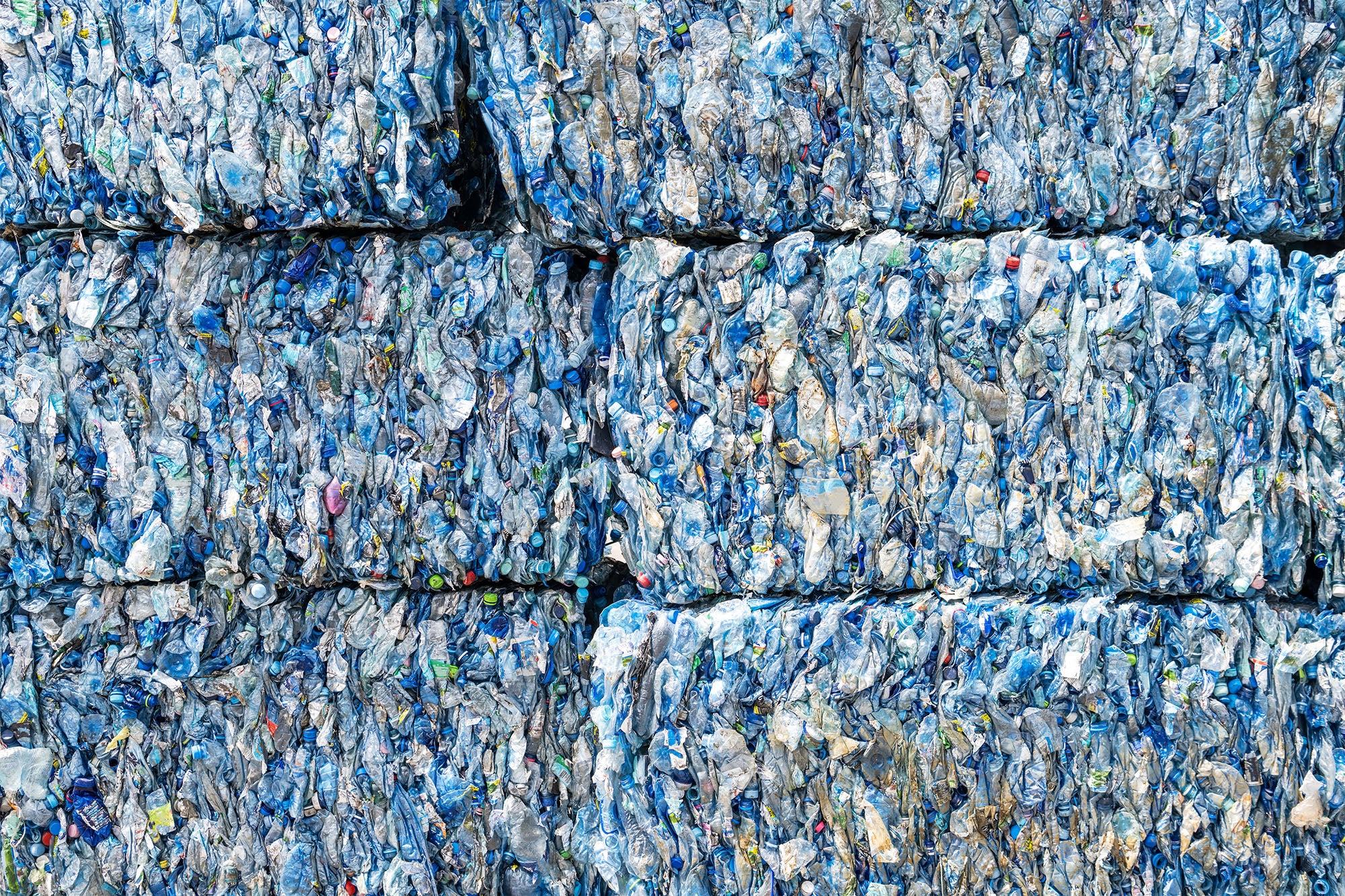 Stacks of tightly compacted, crushed plastic bottles are arranged in large rectangular bales, ready for recycling. The bales are densely packed and display various shades of blue, white, and hints of other colors.