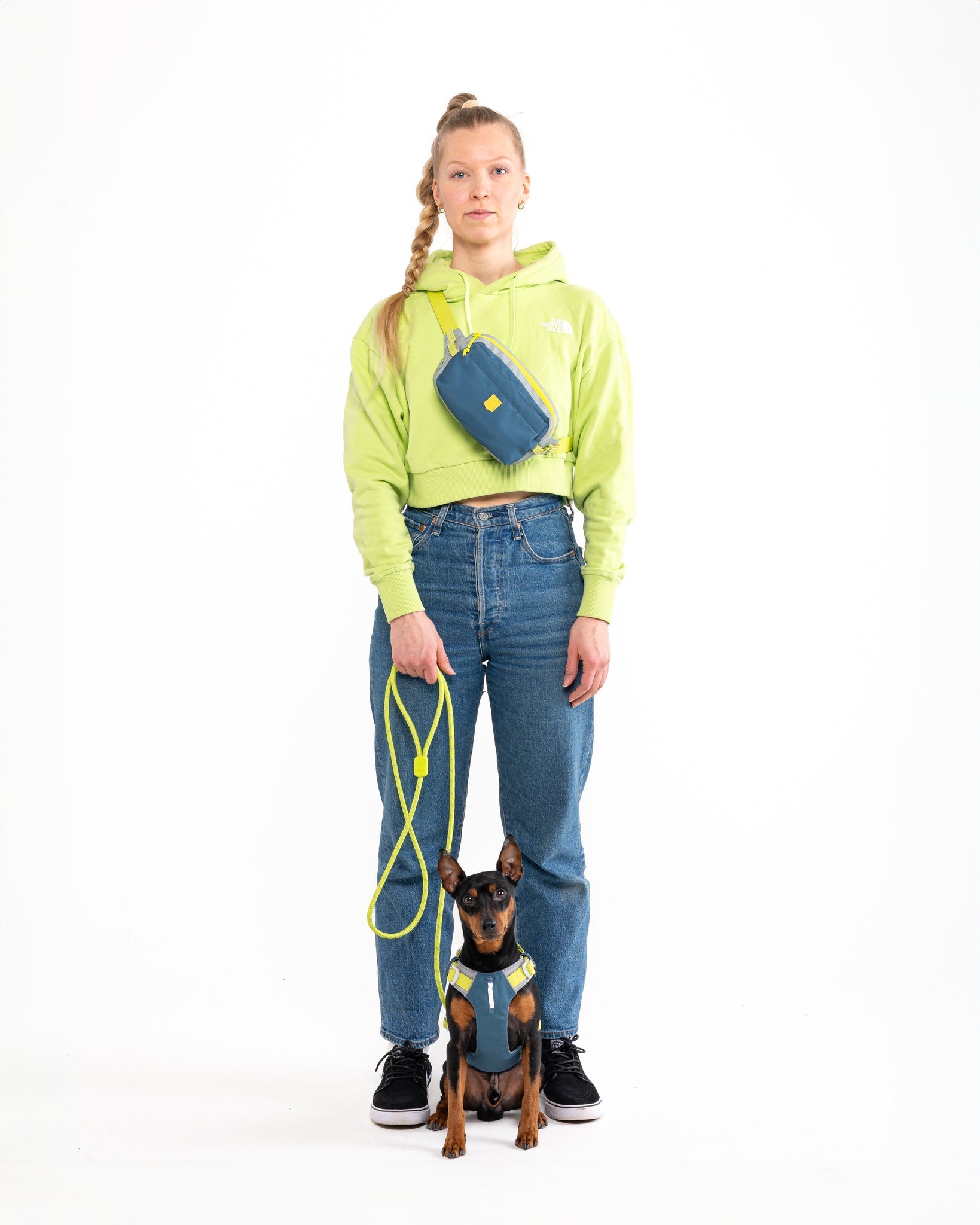A woman in a neon green hoodie, blue jeans, and sneakers stands with a small dog on a neon leash. The dog wears a blue and yellow harness, and the woman carries the Alpha 360 Hip Pack in Deep Teal across her body.