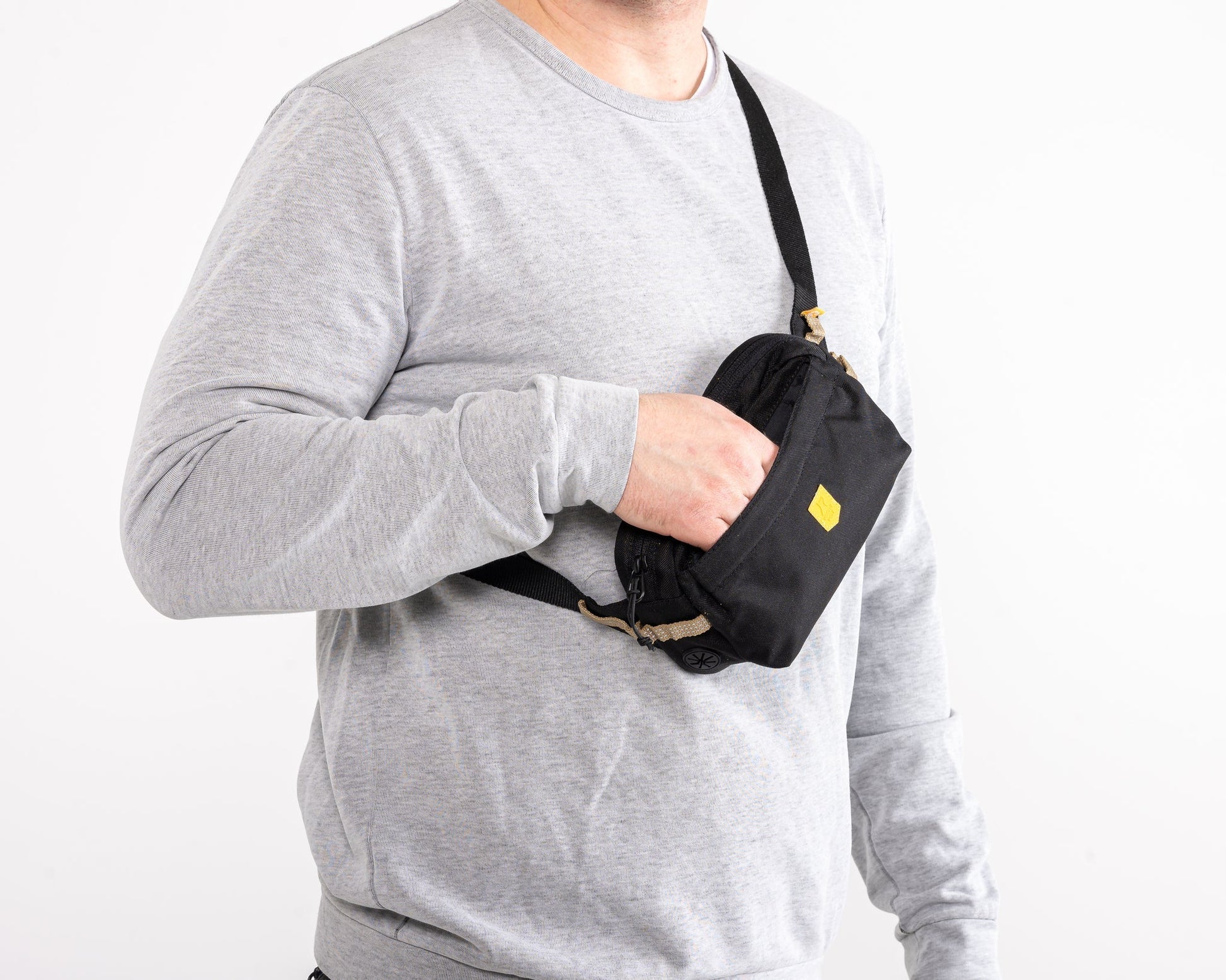 A person in a light gray long-sleeve shirt holds the Alpha 360 Hip Pack Black with a yellow logo, wearing its strap across their torso. The background is plain white.