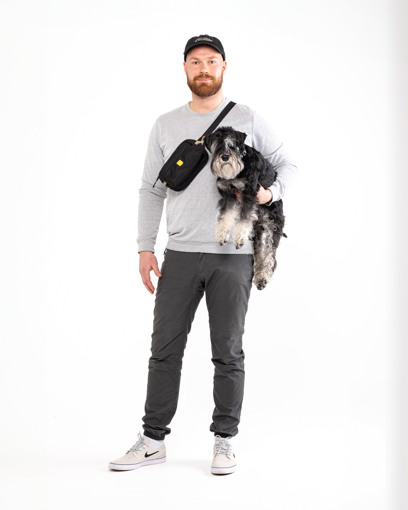 A man stands against a white background, wearing a gray sweatshirt, dark pants, white sneakers, and a black cap. He holds a black and gray dog with one arm and has the Alpha 360 Hip Pack Black crossbody bag slung over his shoulder.