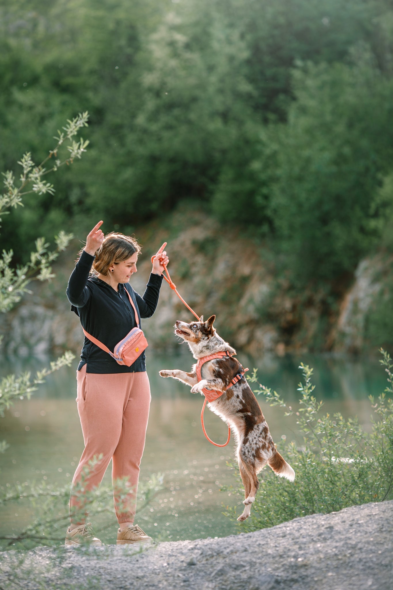 A woman wearing a black sweater and pink Alpha 360 Hip Pack holds a leash as her brown and white dog jumps beside her outdoors, with greenery and water in the background.