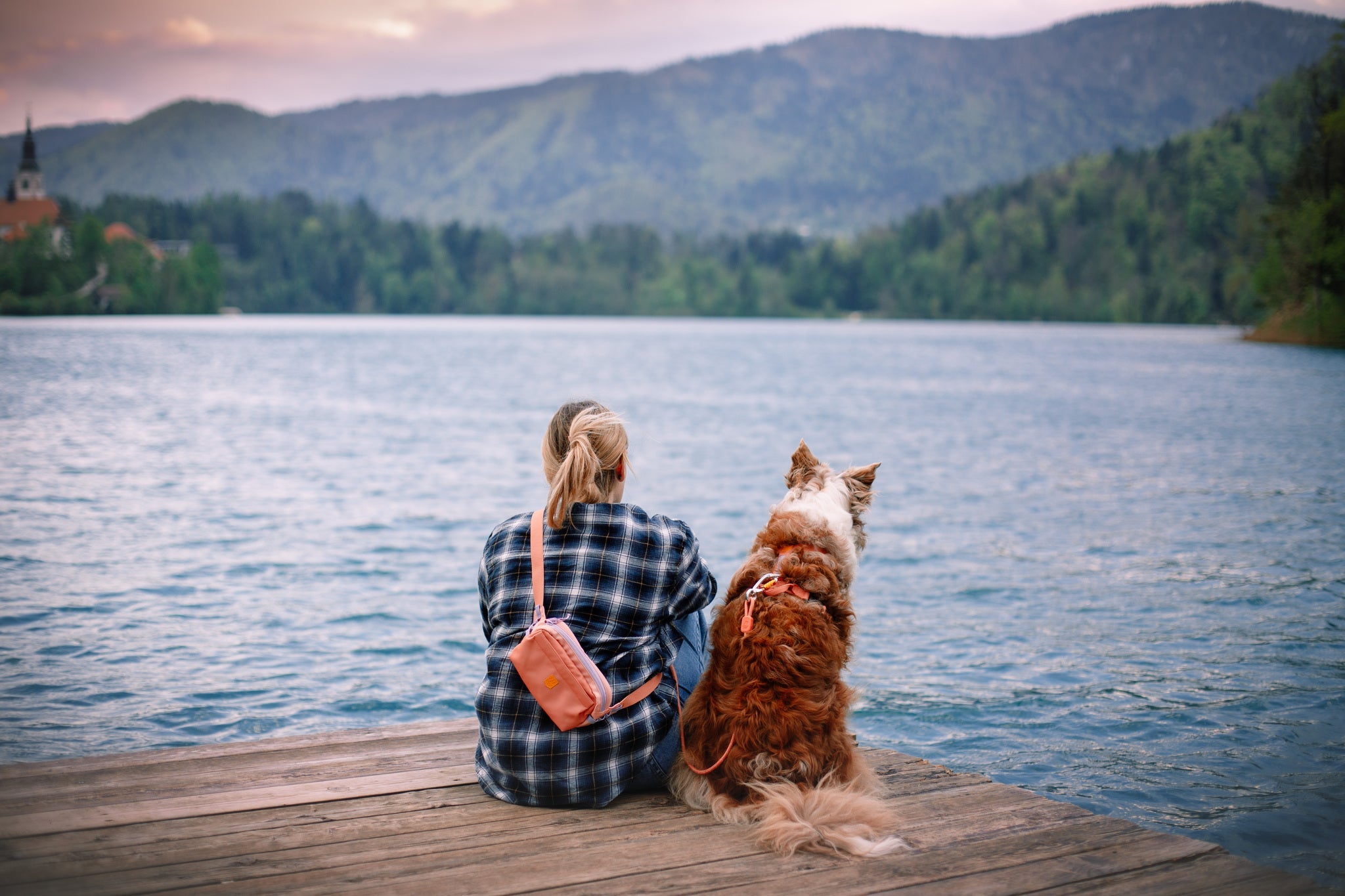 A person with an Alpha 360 Hip Pack in Salmon Pink sits on a wooden dock beside a brown and white dog, both gazing at a calm lake surrounded by forested hills and distant mountains.