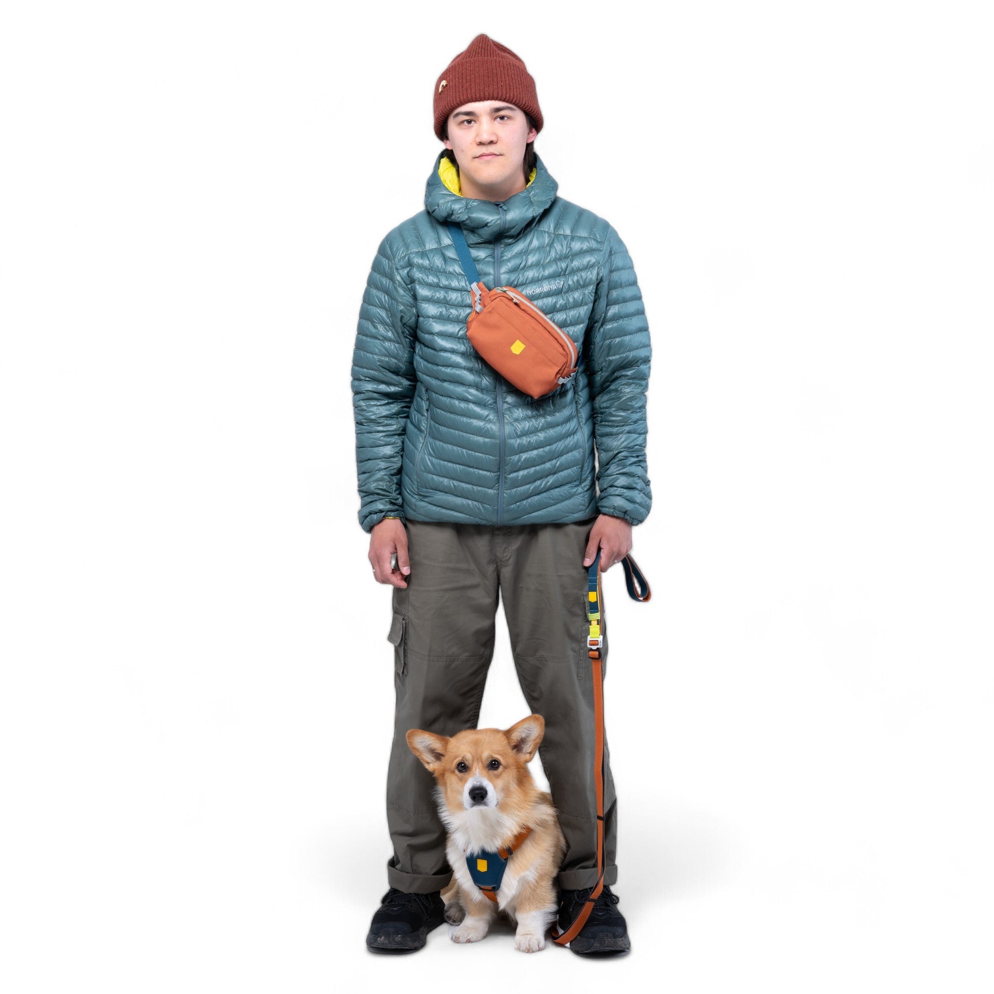 Wearing a teal puffer jacket, brown beanie, and khaki pants, this person holds a leash as their small brown and white dog sits at their feet. They sport the Alpha 360 Hip Pack in Terracotta Mix across their body against a white background.
