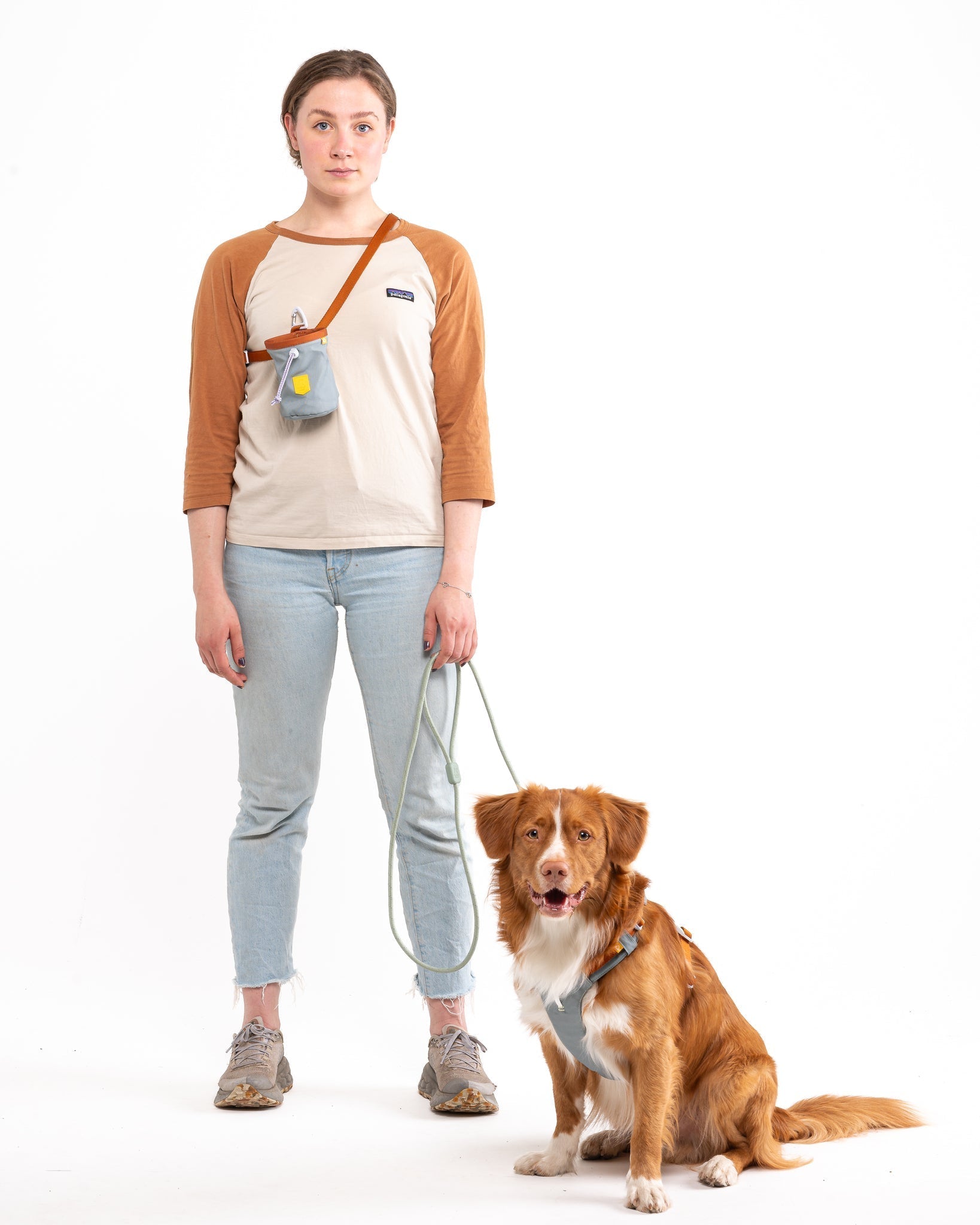 A person in jeans, sneakers, and a beige-orange long-sleeve shirt stands with a leashed brown dog featuring white markings. Both face forward against a plain white background. The Alpha 360 Dog Snack Bag Glacier Green is shown.