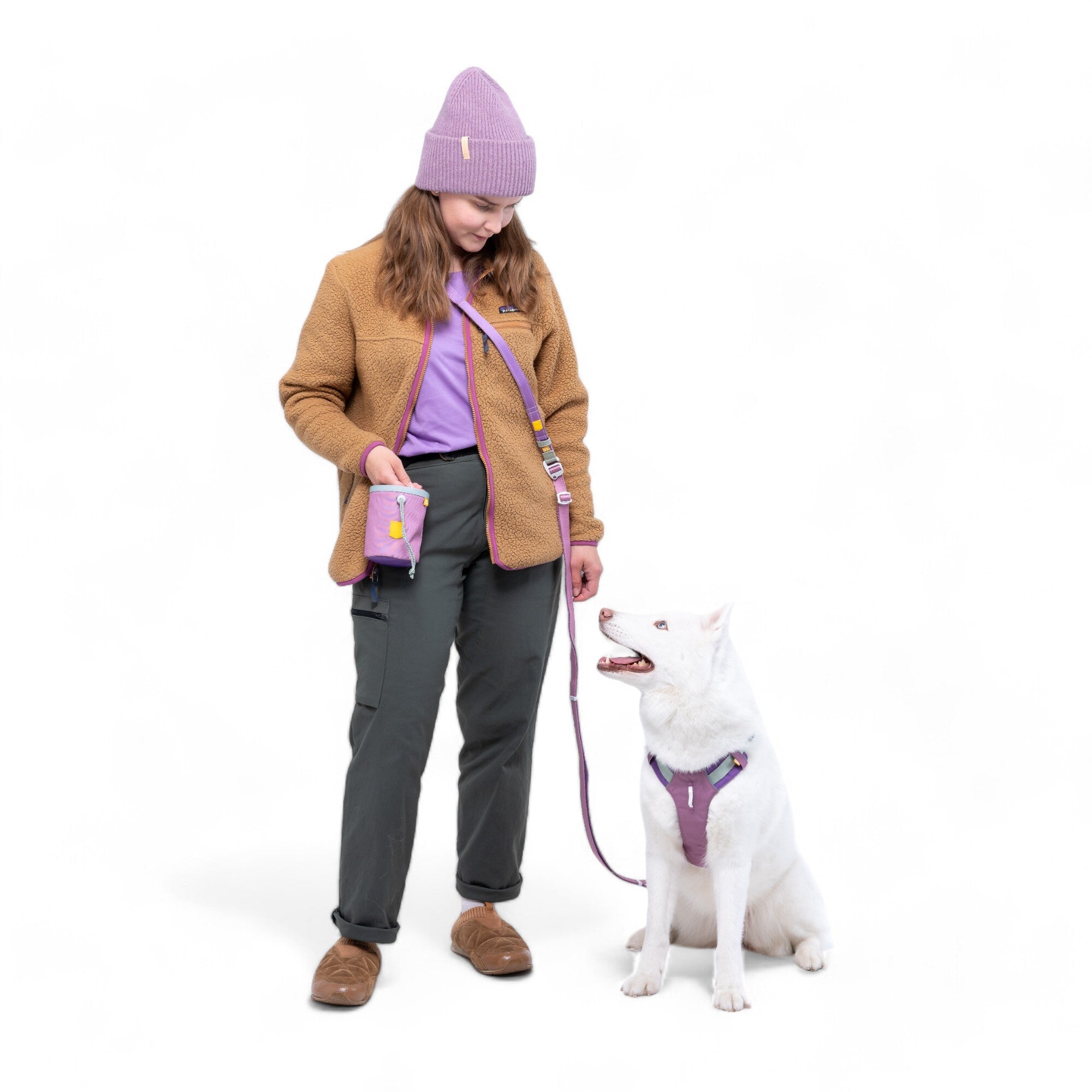A woman in a purple beanie, tan jacket, and gray pants stands on a white background holding the leash of a white dog in a purple harness. She uses the Alpha 360 Dog Snack Bag Mauve Mix while they look at each other.