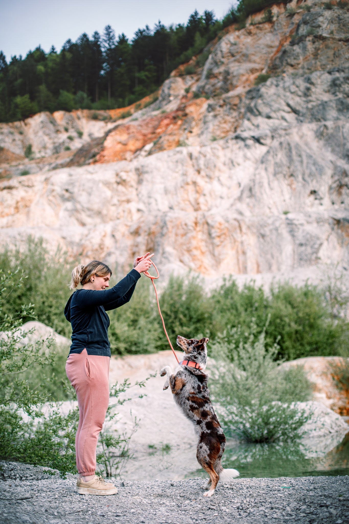 A woman in pink sweatpants holds up a leash attached to a Color Block Martingale Dog Collar Salmon Pink as her spotted dog stands on its hind legs, with rocky hills and green bushes in the background.