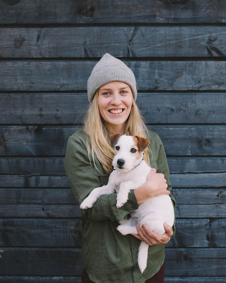 A smiling person in a green shirt and gray beanie holds a small white and brown dog in front of a dark wooden wall.