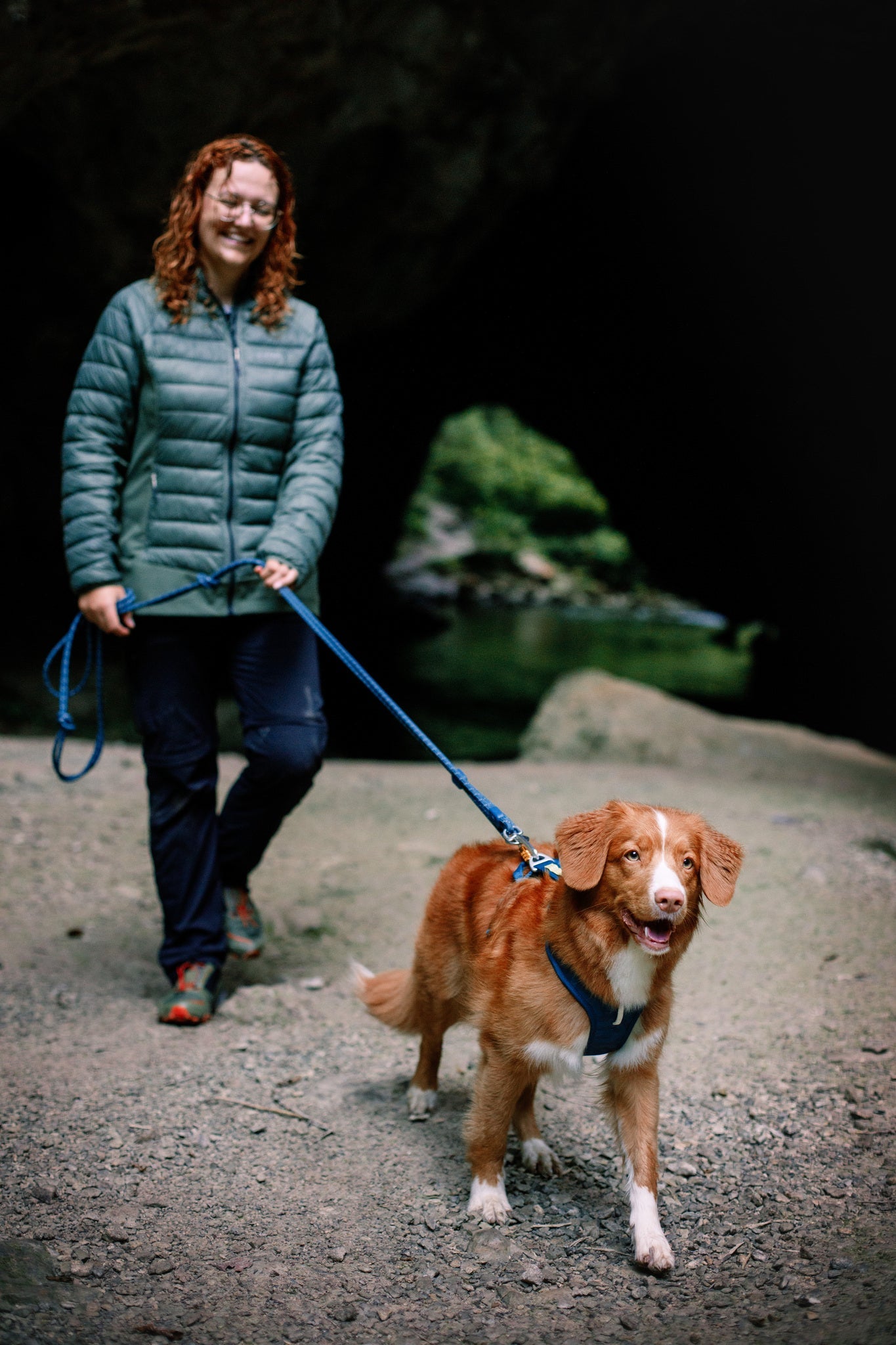 A woman with curly red hair in a green jacket stands on a rocky path, smiling as she holds the leash of her brown and white dog wearing the Roam Dog Harness in Deep Teal. A dark cave entrance is seen in the background.
