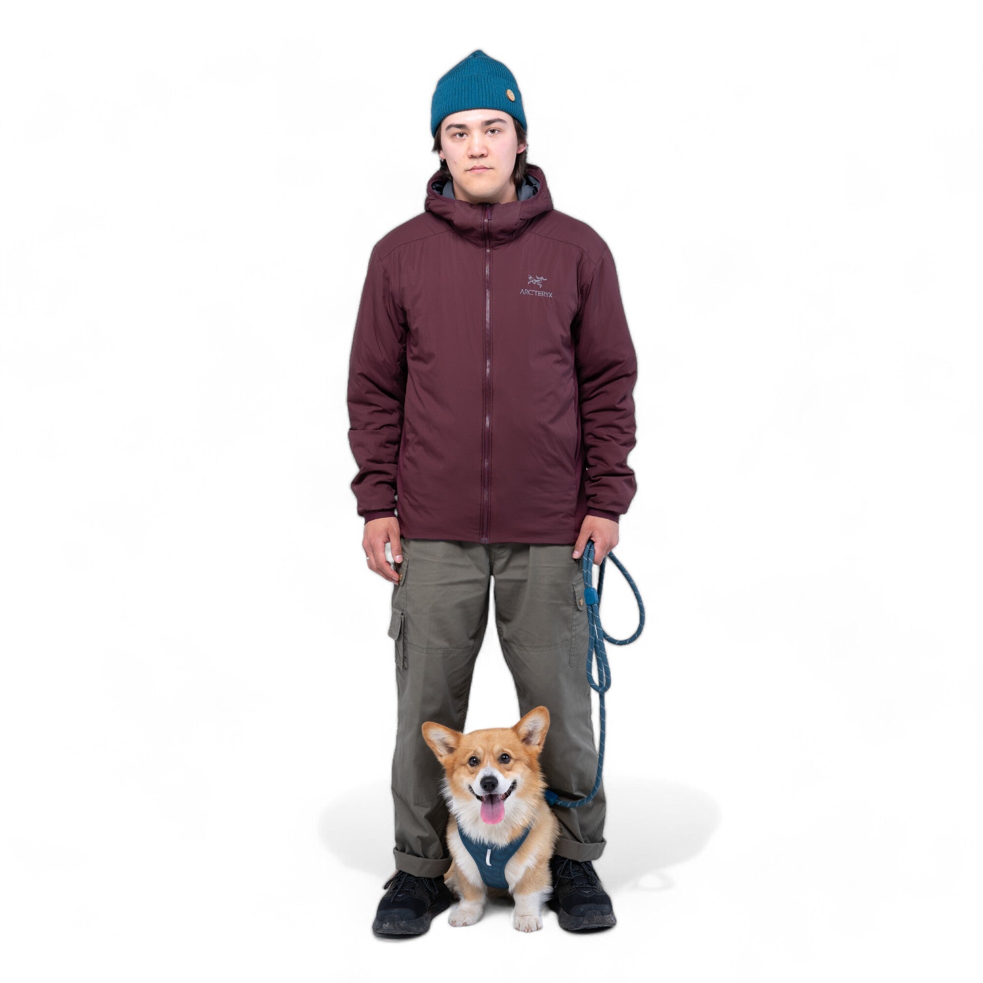A person in a blue beanie, maroon jacket, and cargo pants holds a Rope Dog Leash Deep Teal, with a small corgi sitting in front of them against a white background.
