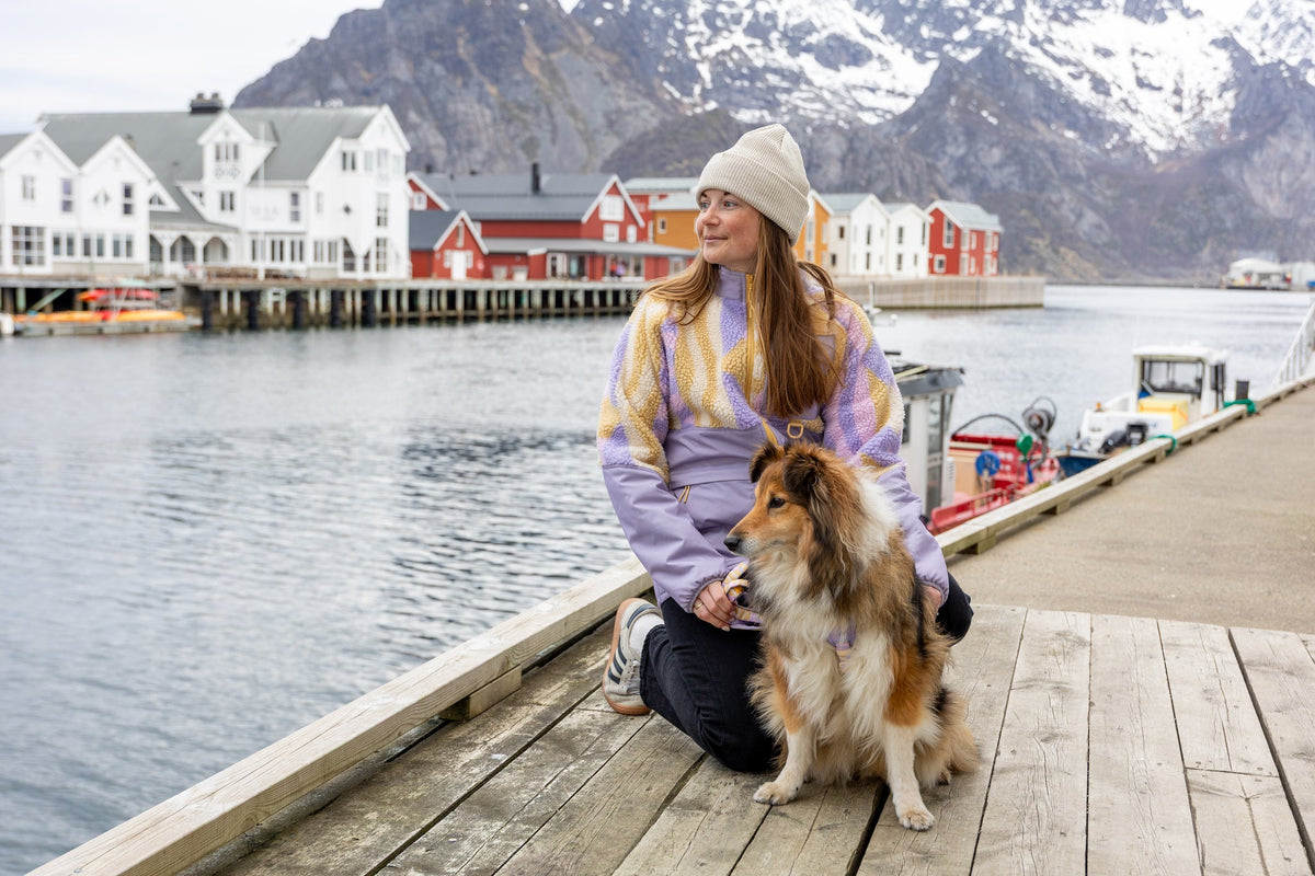 A woman in a knit sweater and beanie sits on a wooden dock by the water, petting a Collie dog. Snowy mountains and colorful houses are visible in the background.