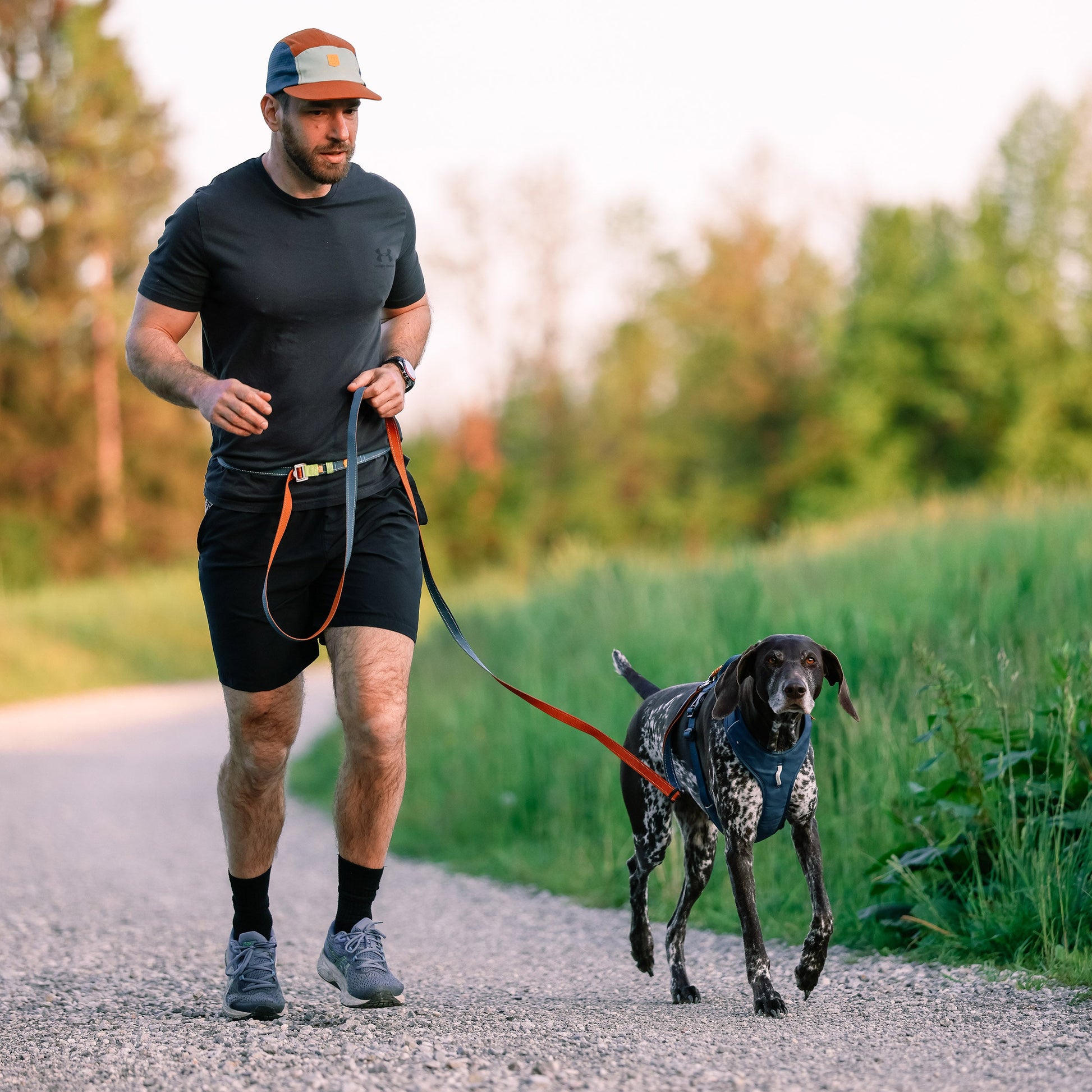Man jogging on a gravel path with a black and white dog on a leash, surrounded by greenery.