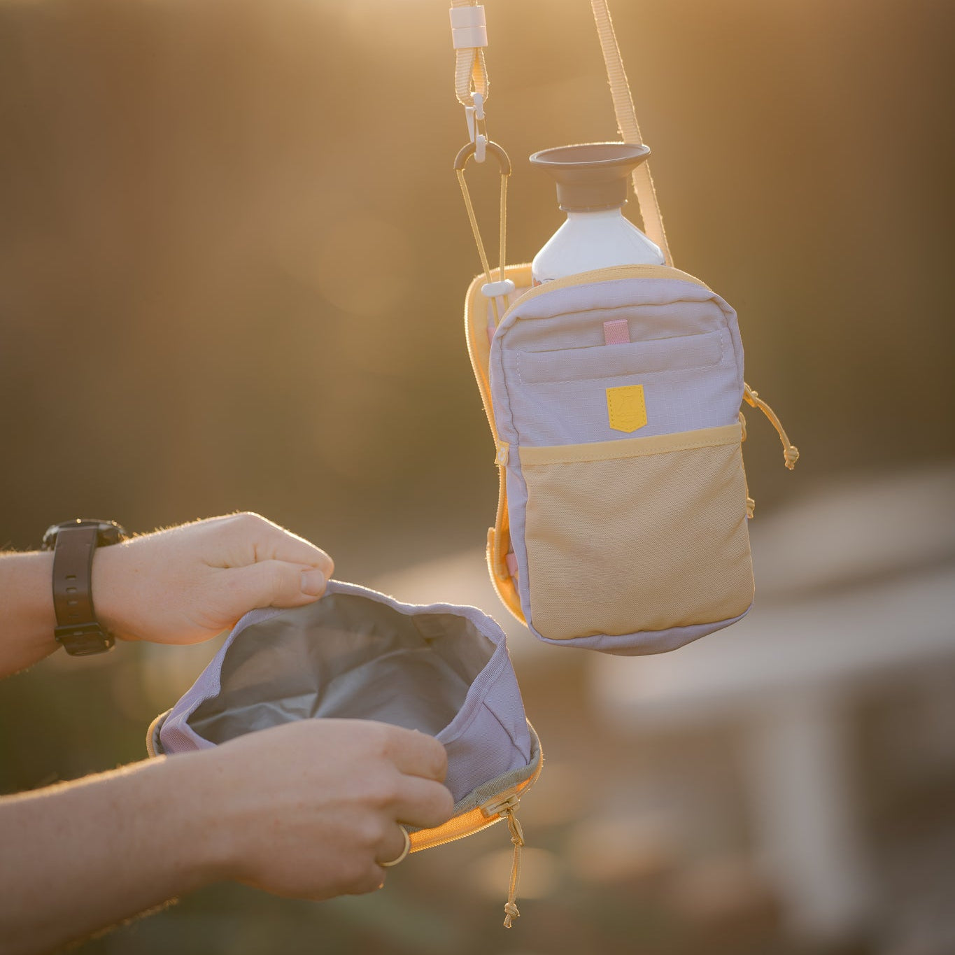 Two small bags, one hanging with a water bottle, another held open by a person, outdoors at sunset.