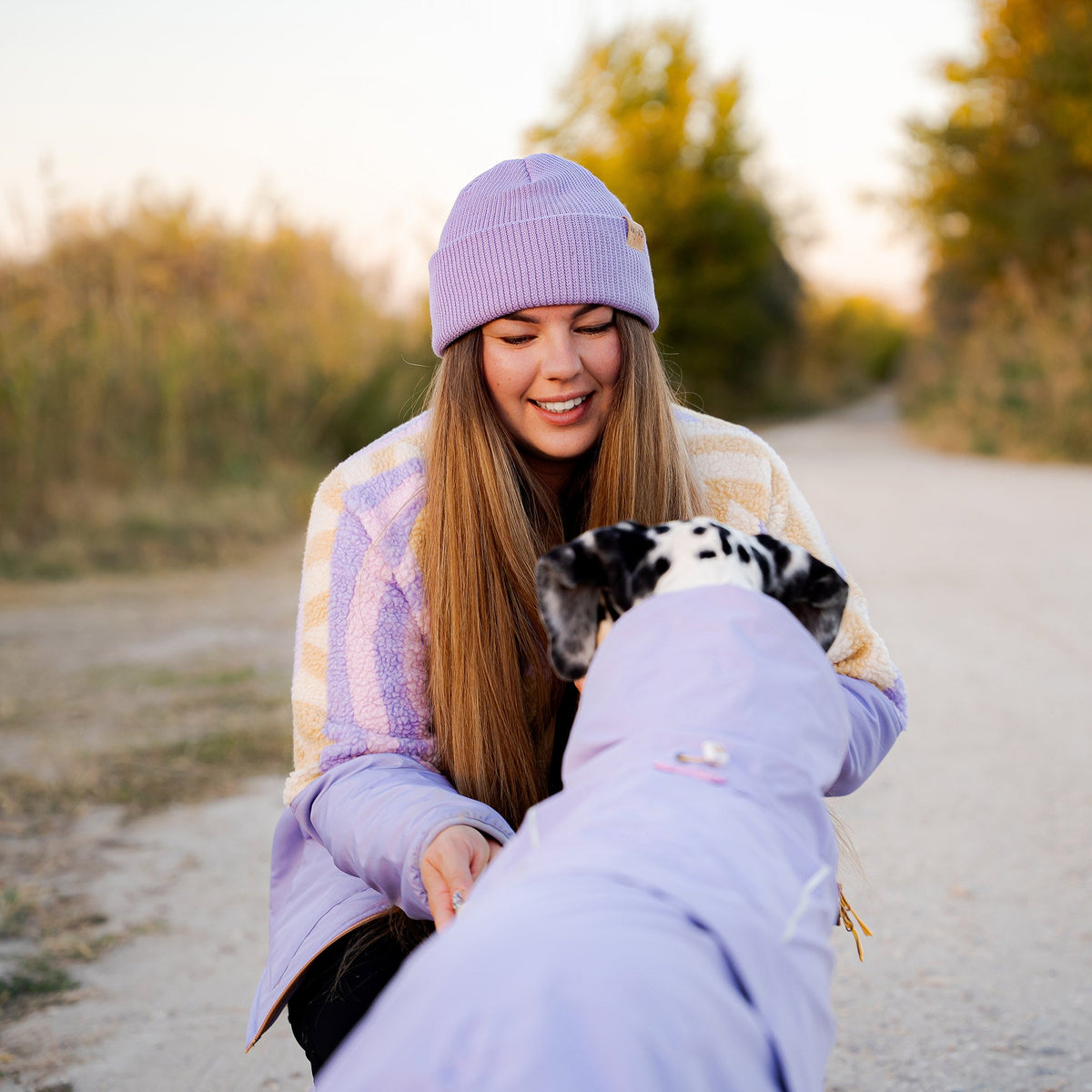 A smiling woman wearing a lilac beanie and jacket interacts with a dalmatian dog on a dirt path.