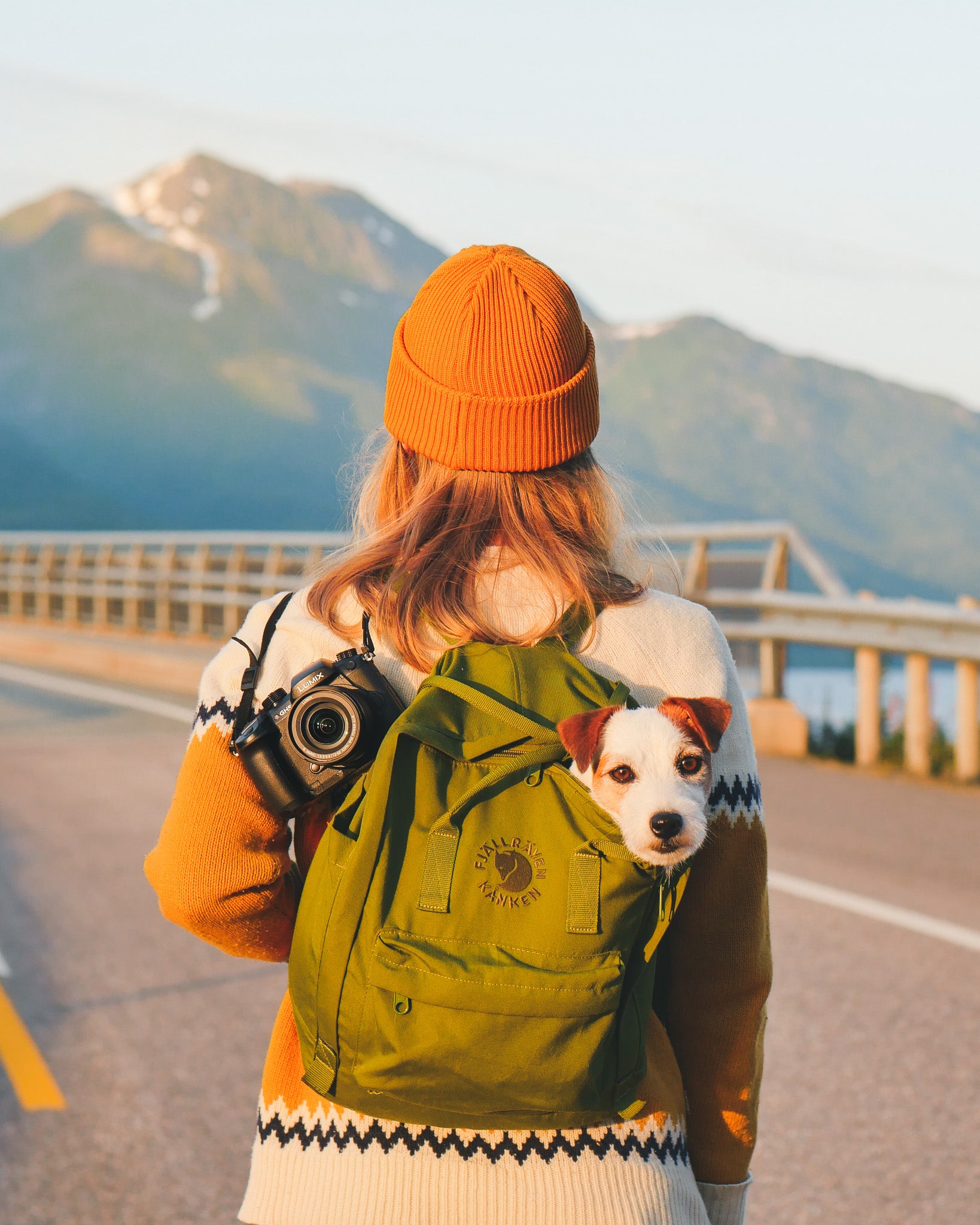A person wearing an orange beanie and sweater stands on a road with mountains in the background. They have a green backpack with a small dog peeking out and a camera hanging from their shoulder.