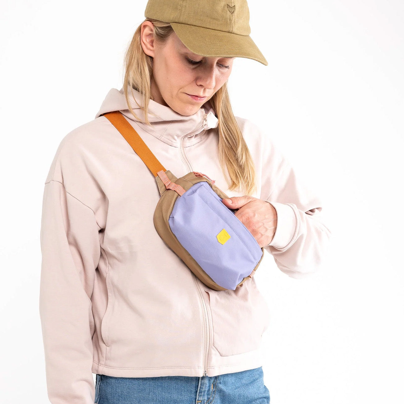 Woman in a beige cap and light jacket adjusts a small purple and tan crossbody bag against a white background.